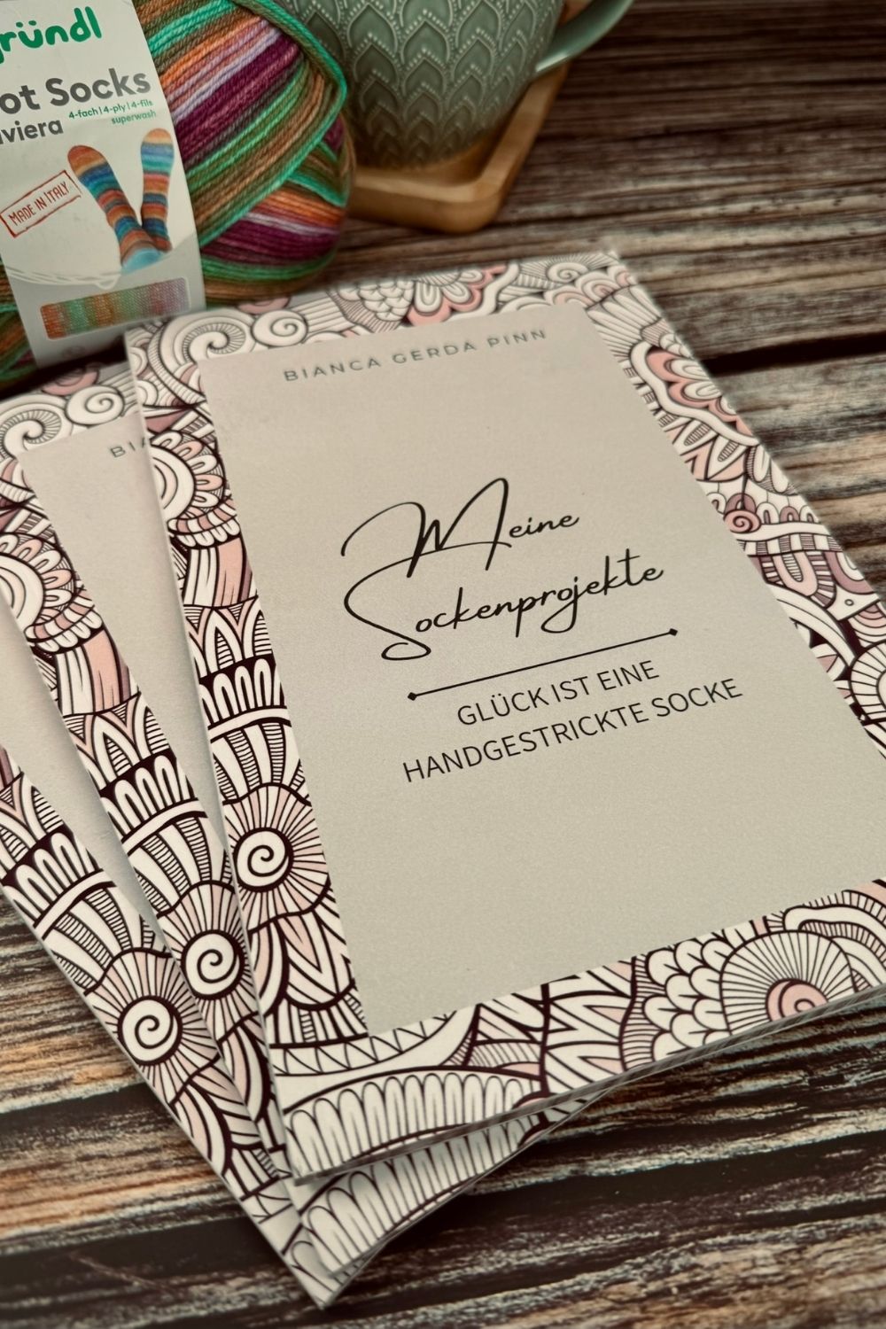 Drei Bücher "Meine Sockenprojekte" auf einem Tisch aufgefächert, im Hintergrund eine Tasse Tee und Sockenwolle