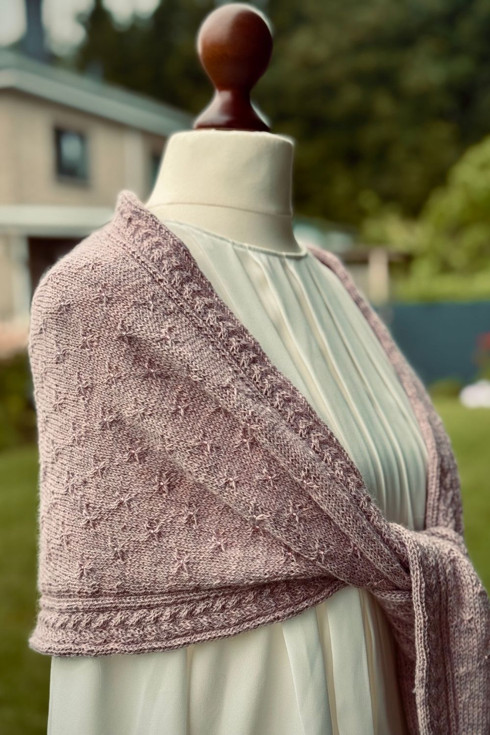 Das Tuch Lyra in altrosa kombiniert mit einer weißen Bluse, lässig geknotet auf einer Schneiderpuppe vor weichgezeichnetem Gartenhintergrund