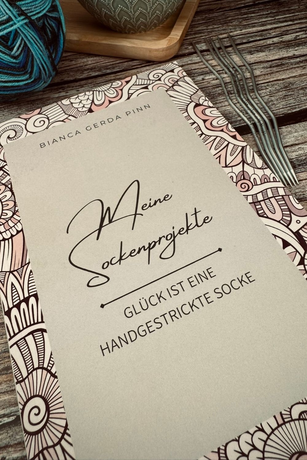 Bücher "Meine Sockenprojekte" gestapelt auf dem Tisch, im Hintergrund eine Tasse Tee, Sockenwolle und Stricknadeln