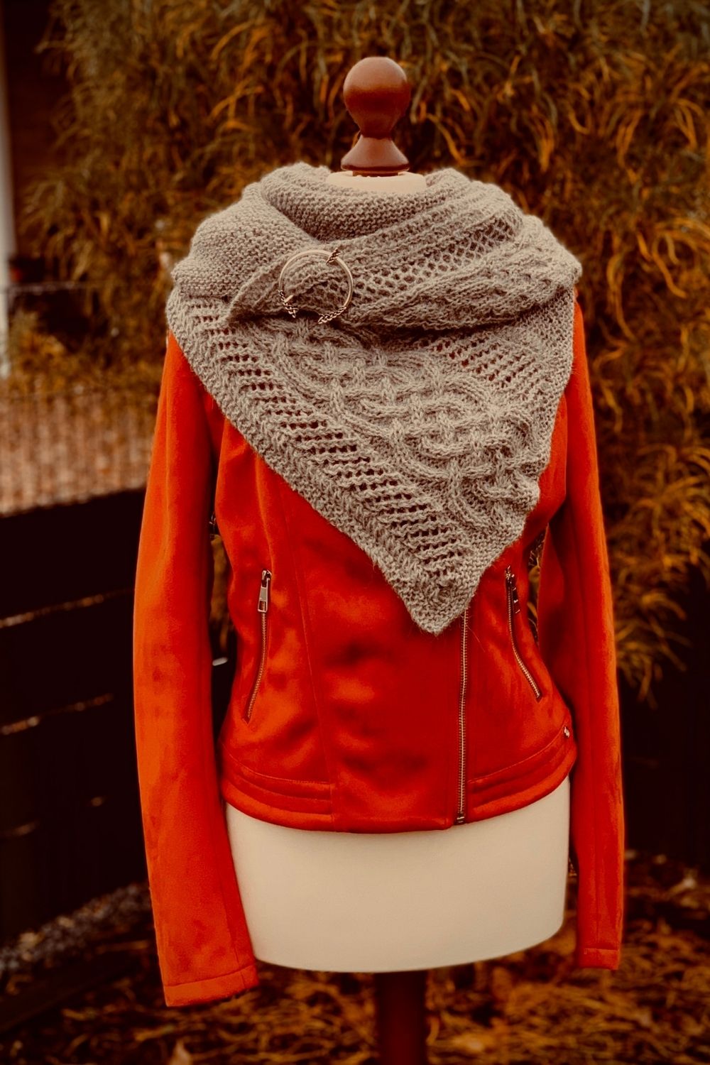 Asymmetrisches Stricktuch Celtic Mystery über einer orangefarbenen Lederjacke auf einer Schneiderpuppe im Garten präsentiert