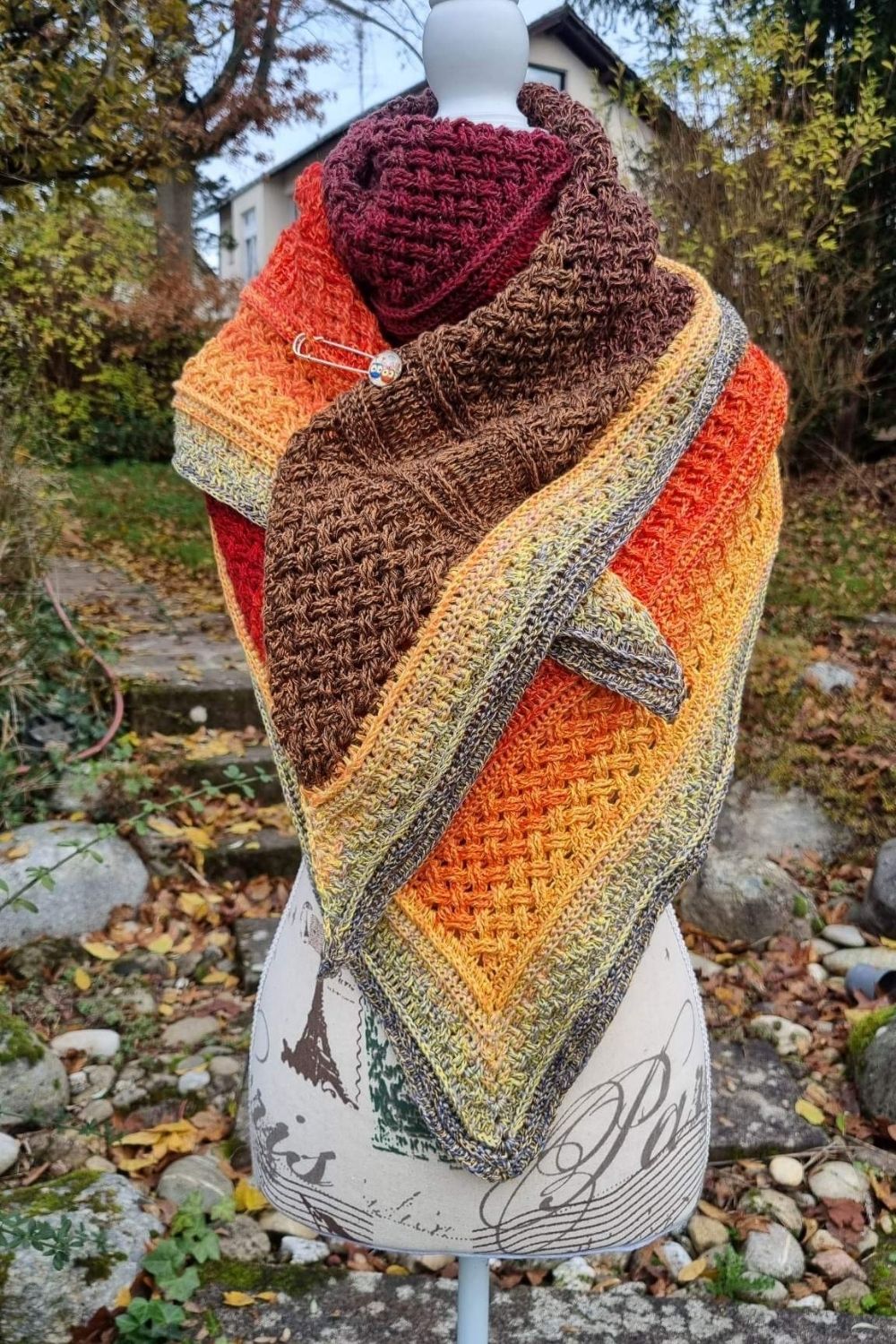 Das Tuch Flechtwerk in herbstlichen Farben, lässig gebunden mit Tuchnadel auf einer Schneiderpuppe vor weichgezeichnetem Gartenhintergrund
